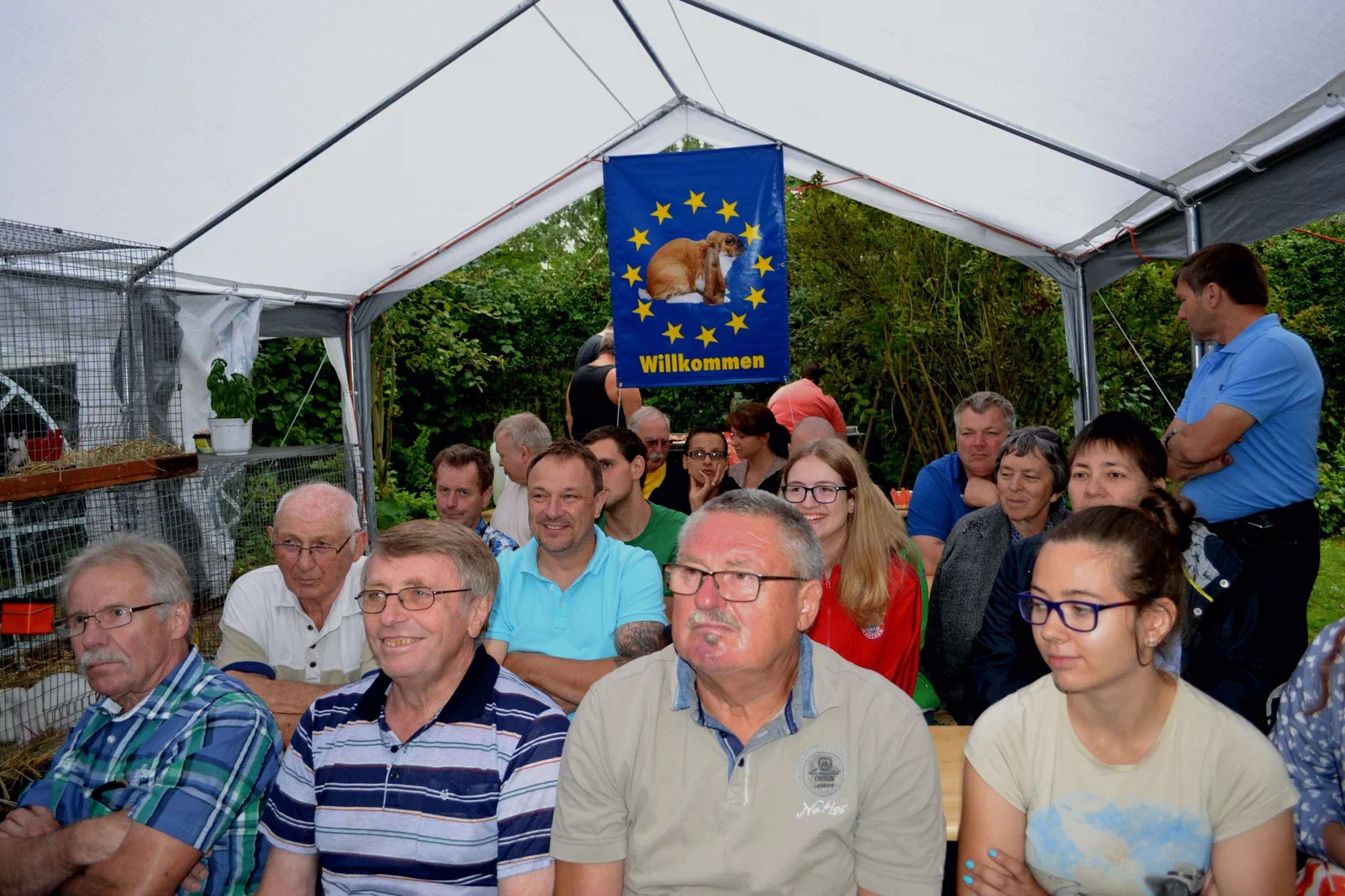 Gäste und Preisrichter sitzen auf Bänken im Zelt, hinter ihnen hängt eine blaue Fahne mit Kaninchenbild und der Aufschrift „Willkommen“