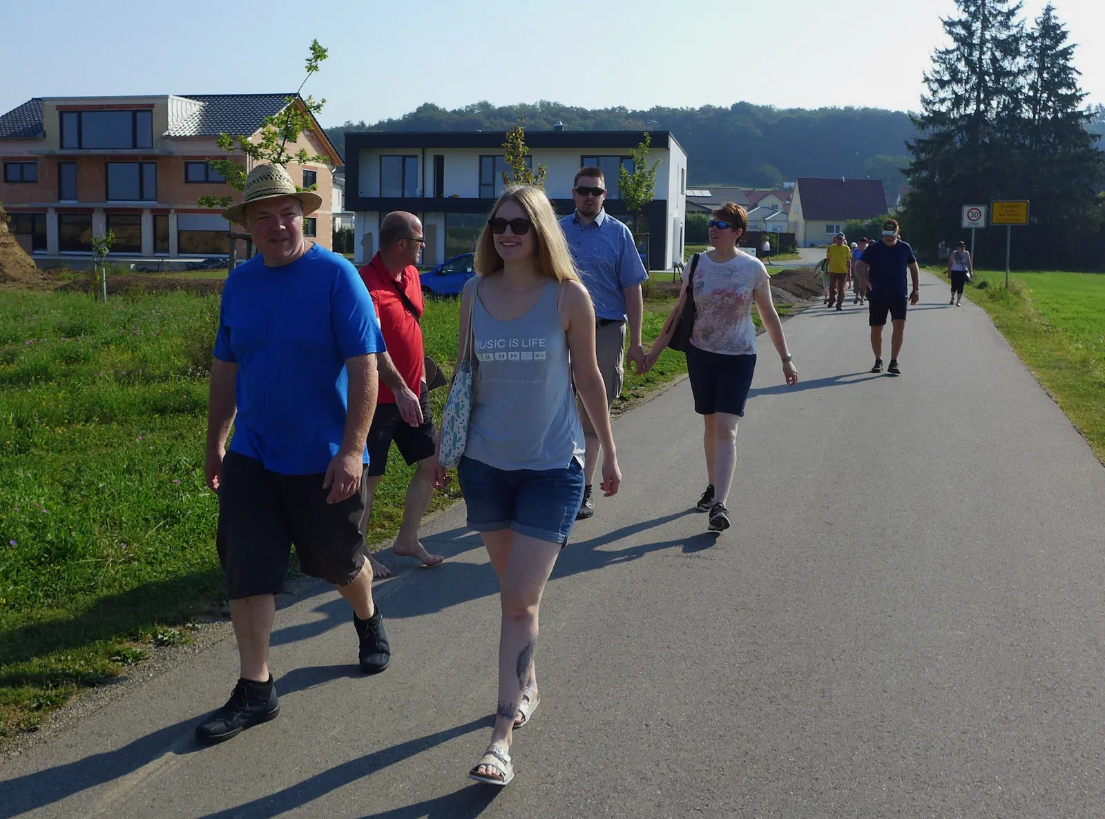Teilnehmer spazieren bei Sonnenschein auf einem Feldweg am Ortsrand, im Hintergrund Neubauten und bewaldete Hügel