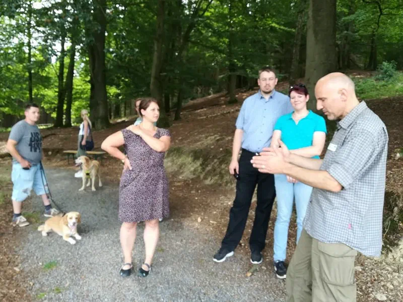 Ein Museumsführer erklärt einer kleinen Gruppe von Teilnehmern etwas auf einem Waldweg, im Hintergrund zwei Hunde