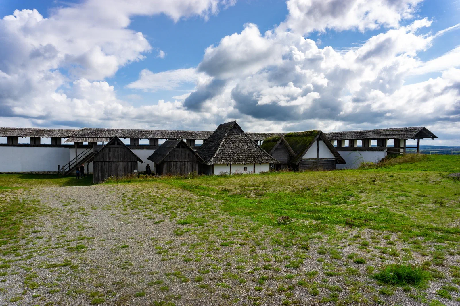 Maisons celtes en bois reconstituées, aux toits à deux pans, devant un mur d&rsquo;enceinte crépi de blanc avec un chemin de ronde couvert, devant une prairie sous un ciel nuageux