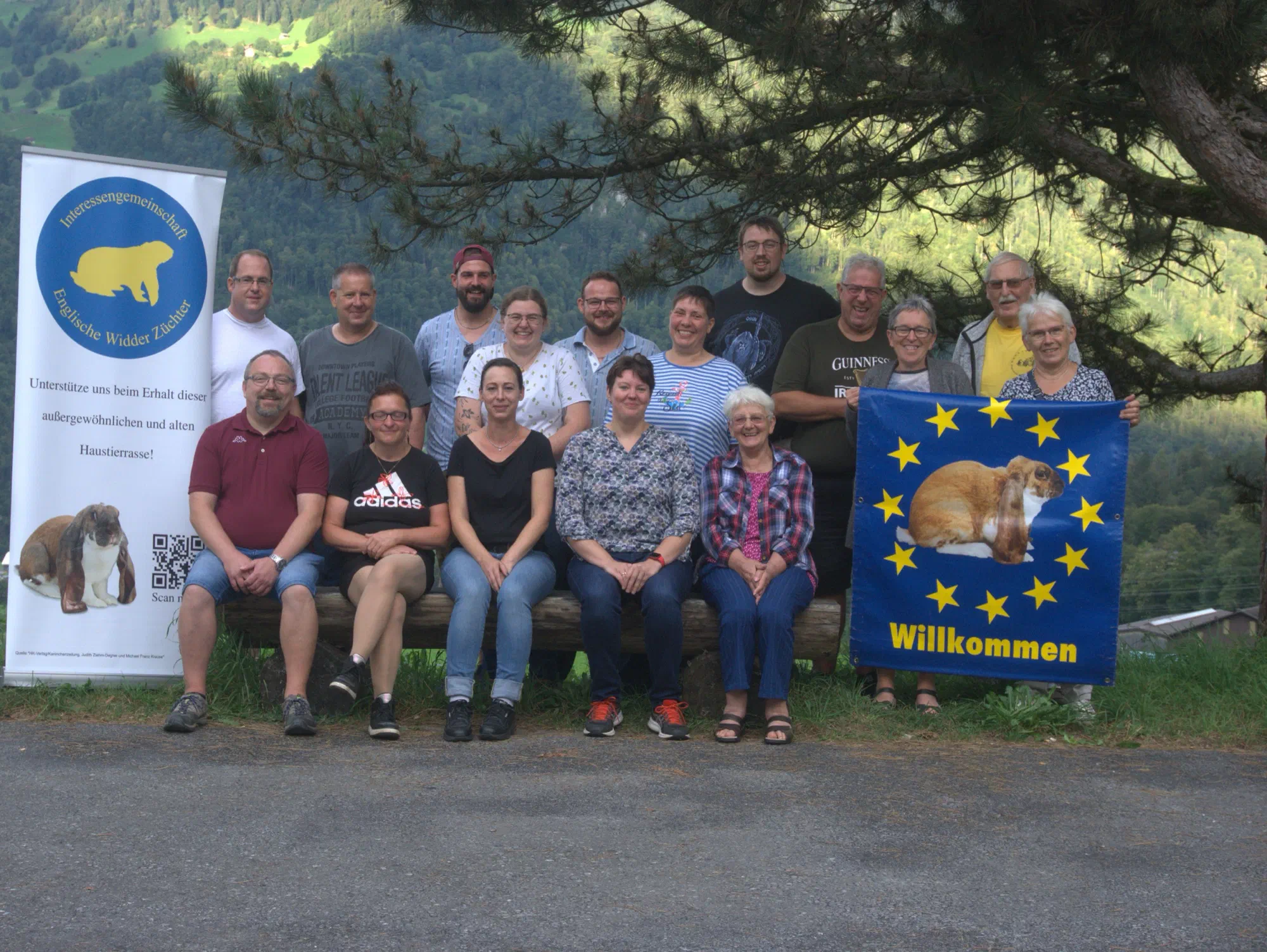 Photo de groupe des participants devant une forêt de montagne, à droite un drapeau bleu avec une couronne d'étoiles, un lapin et l'inscription Willkommen (bienvenue), à gauche une banderole enroulable de l'IG