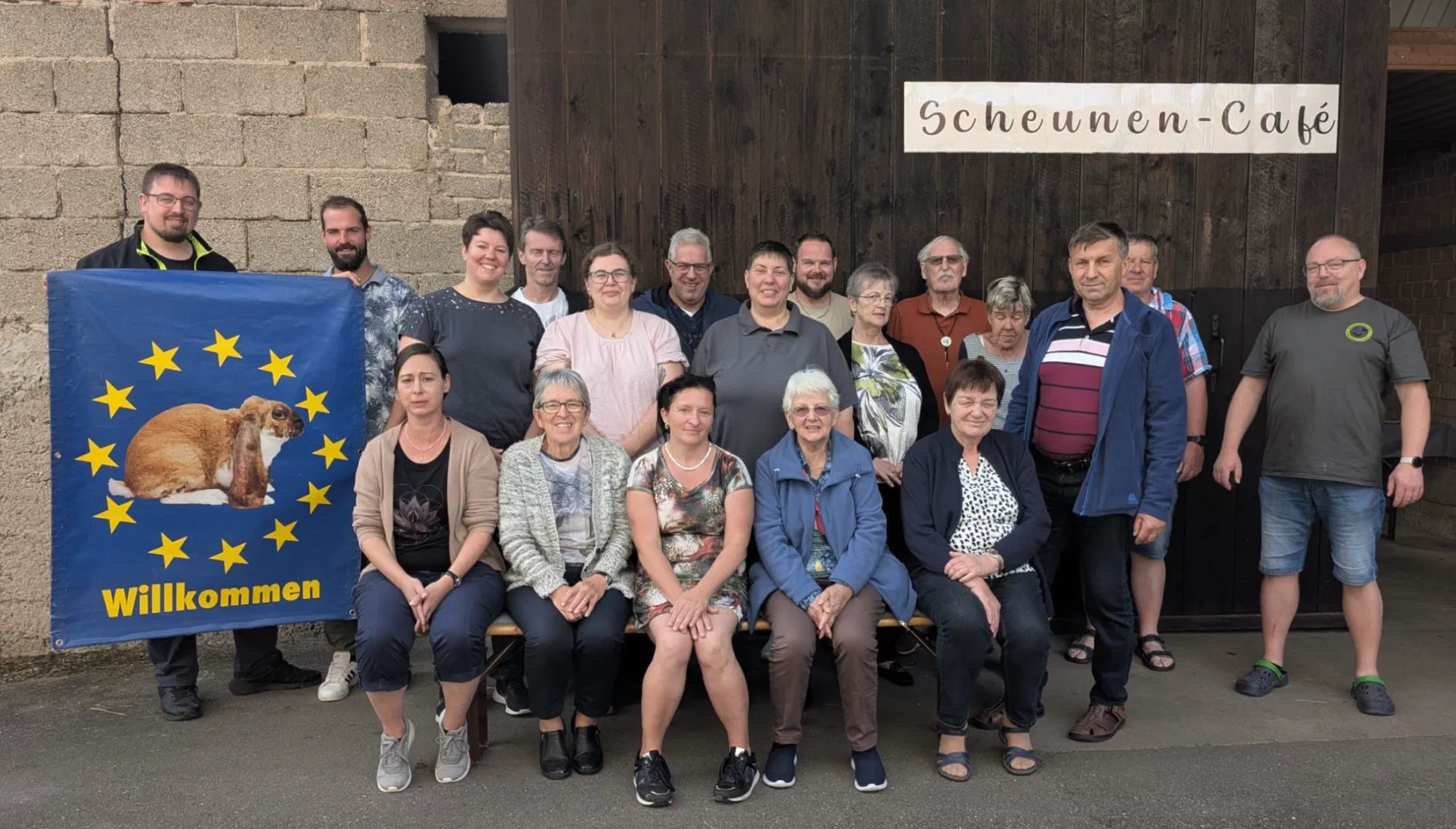 Photo de groupe des participants devant une grange portant l'enseigne Scheunen-Café, à gauche un drapeau bleu avec une couronne d'étoiles, un lapin et l'inscription Willkommen (bienvenue)
